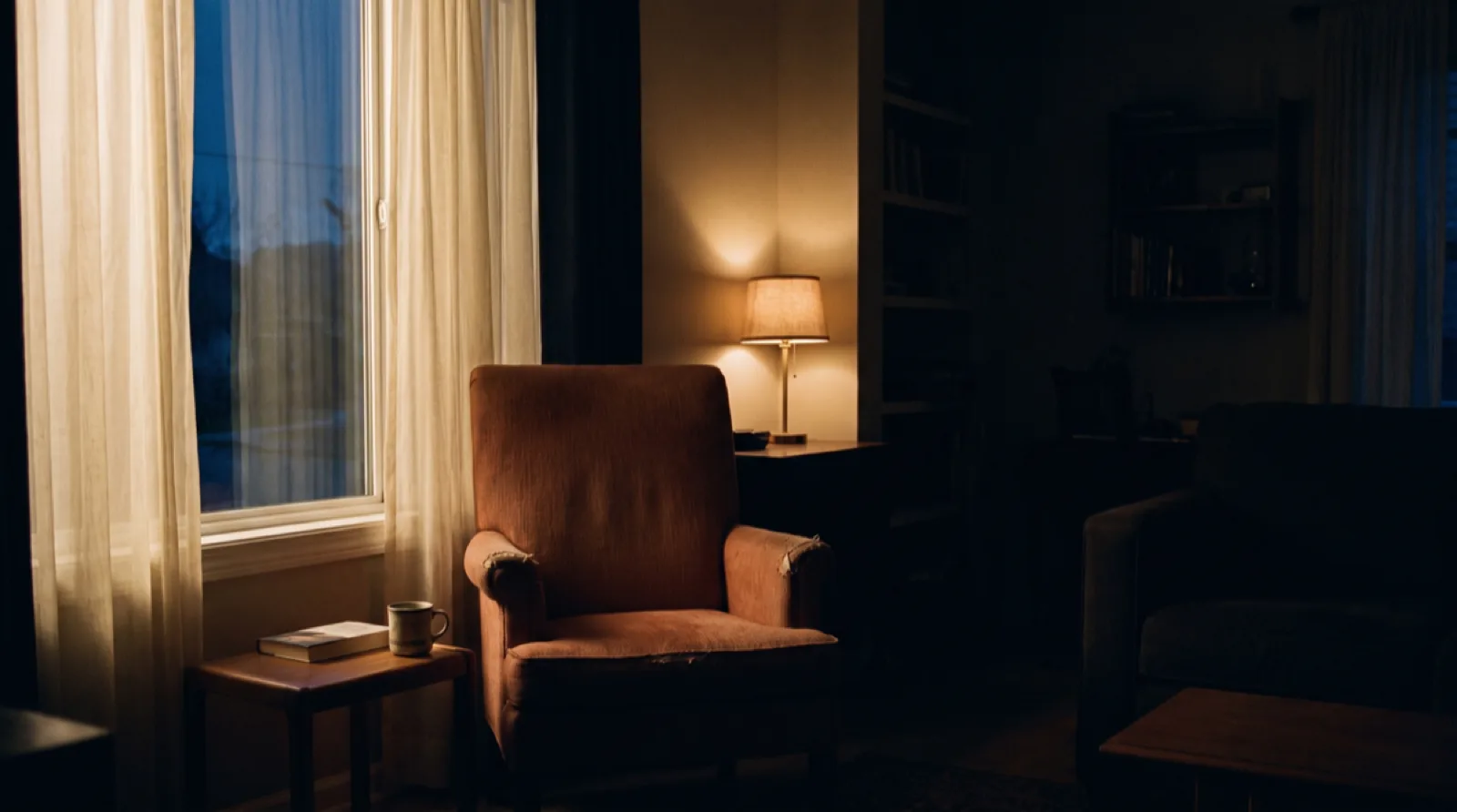 Un fauteuil près d'une fenêtre au crépuscule, une lampe allumée au fond